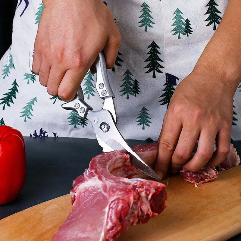 Wearing a tree-patterned apron, a person uses OWOW Me Stainless Steel Poultry Bone Cutter Scissors to cut raw meat on a wooden cutting board, with a red bell pepper beside them.