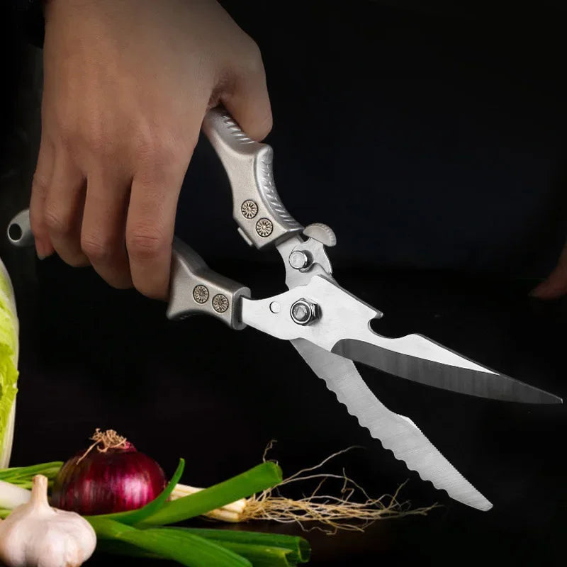 A hand holds the OWOW Me Stainless Steel Poultry Bone Cutter Scissors above green onions, red onion, garlic, and leafy greens on a dark background.