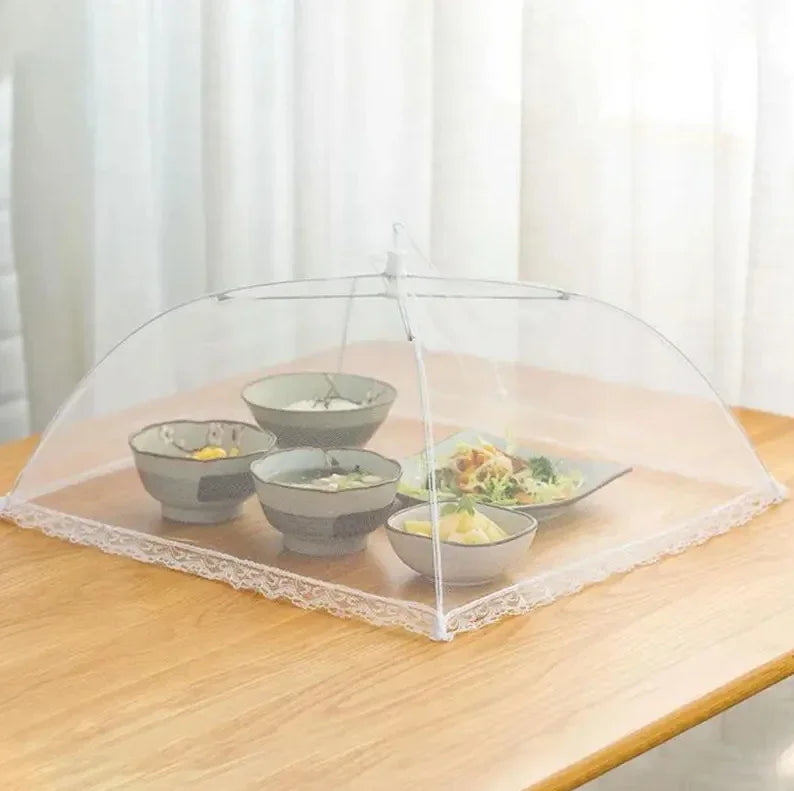 Four bowls of food and a salad plate rest on a wooden table, covered by the OWOW Me Best foldable anti-fly screen mesh for food in white, with lace trim, against a light-colored curtain background.
