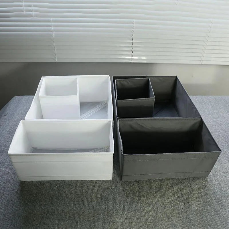 Two OWOW Me Large foldable clothes organizers with compartment storage boxes, in white and gray, are shown side by side on a gray surface with window blinds in the background.
