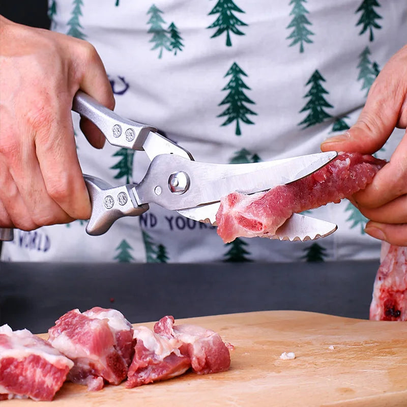 A person uses OWOW Me Stainless Steel Poultry Bone Cutter Scissors to cut raw meat on a wooden cutting board while wearing an apron with a green pine tree pattern.